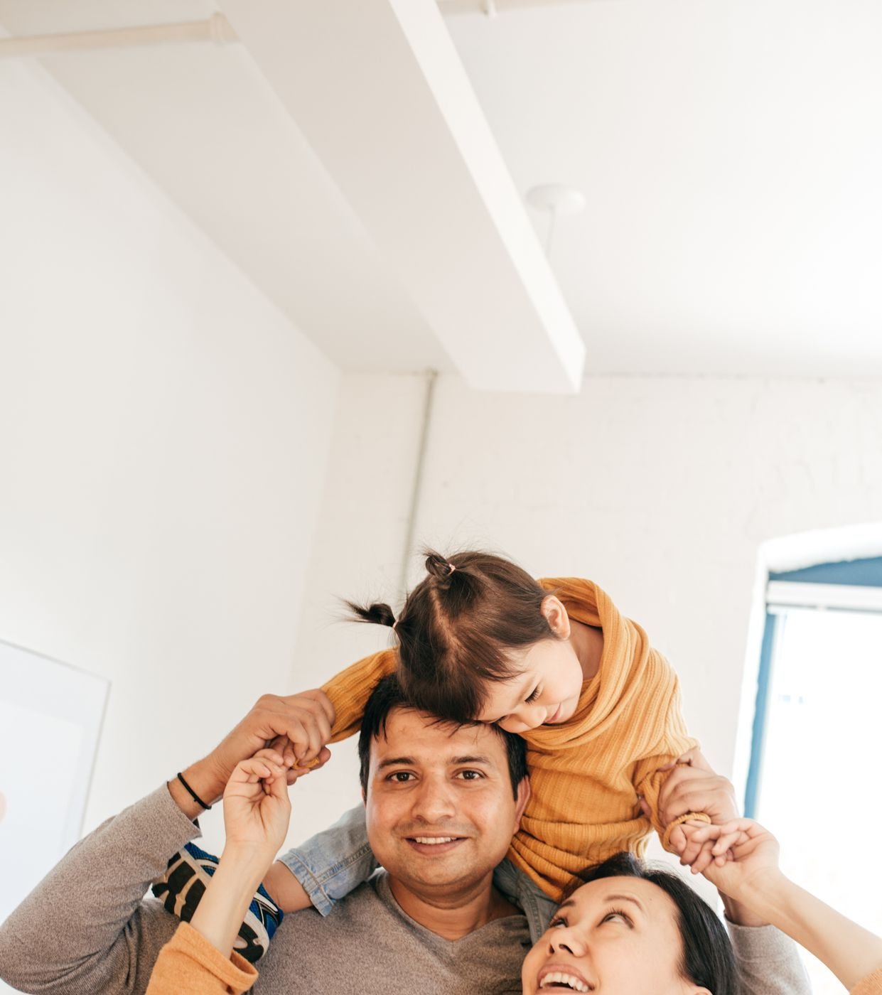 A smiling family: a child on her father's shoulders while her mother looks up and laughs.