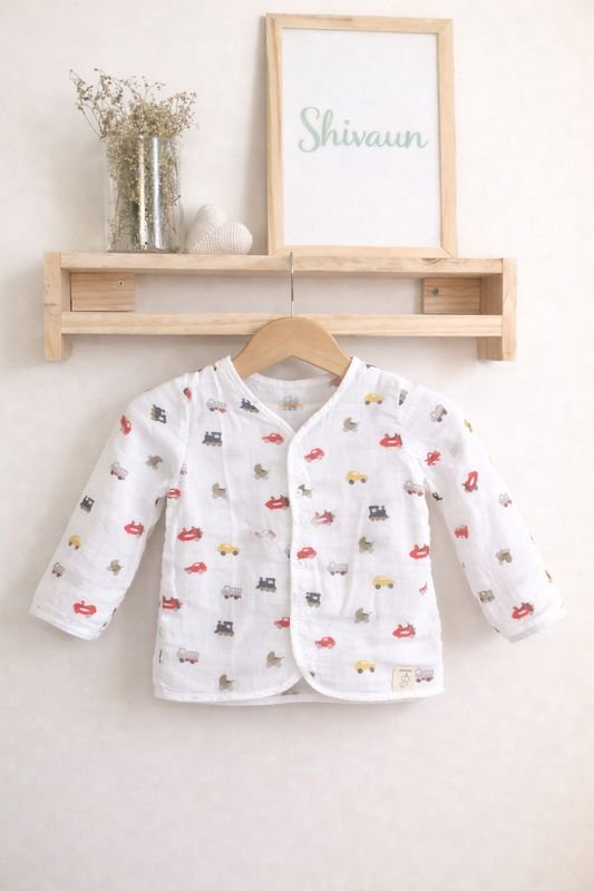 A white muslin baby shirt with a toy-car print on a wooden hanger.