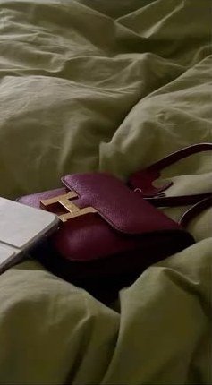A burgundy leather handbag on rumpled sage bedding.