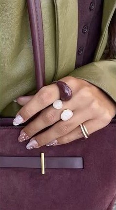 A hand with white stone rings resting on a plum suede bag.