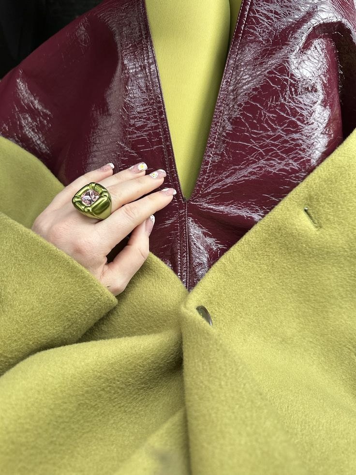 A hand with a green statement ring resting on a chartreuse wool coat over a burgundy patent top.